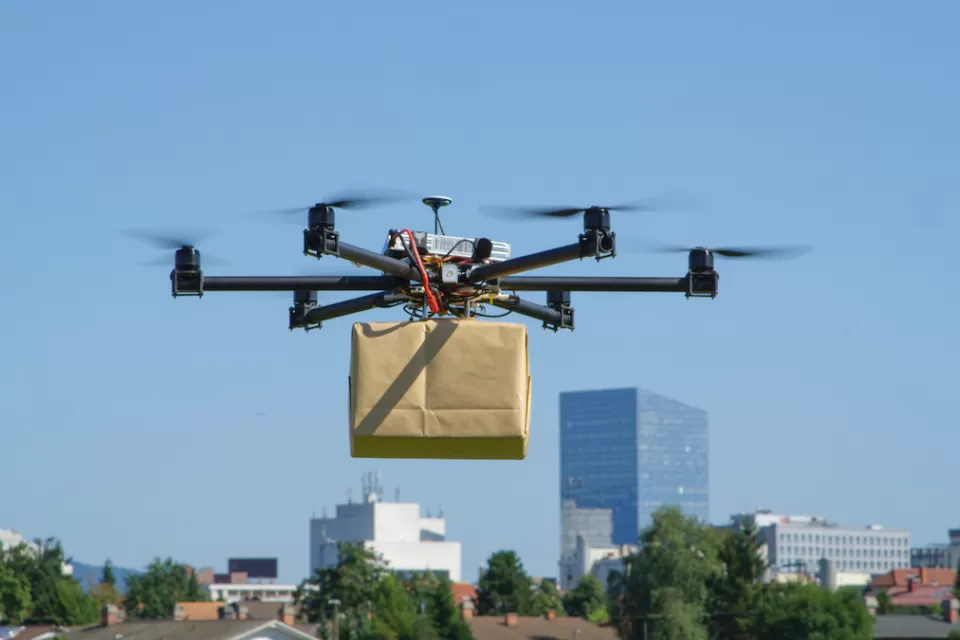 Delivery drone holding a brown paper wrapped box hoveringin air with city in background.
